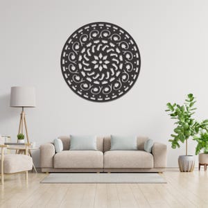 Op de afbeelding: Een groot, rond zwart metalen wandkunstwerk met een ingewikkeld, opengewerkt ontwerp. Het kunstwerk heeft een centraal bloemmotief omgeven door wervelende patronen en decoratieve uitsparingen. Het is gemonteerd op een witte muur boven een beige bank.