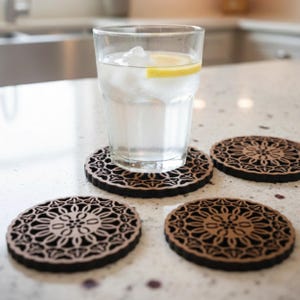 以下が含まれることがあります： 氷とレモンスライスが入った水の入ったグラスが、装飾的な木製のコースターの上に置かれています。花柄のコースターが他に3つ見えます。コースターは茶色で、黒い縁取りがあります。