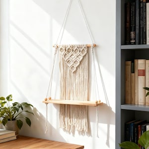 May include: A macrame hanging shelf with a natural wood shelf and a cream-colored woven design. The shelf is suspended by cream-colored cords and is mounted on a white wall. The shelf is approximately 24 inches wide.