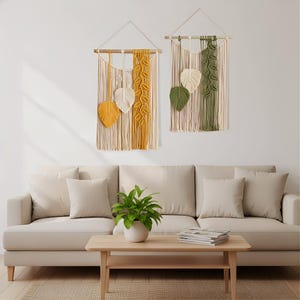 以下が含まれることがあります： 葉の模様が施された2つのマクラメ壁掛け。1つは黄色とクリーム色、もう1つは緑とクリーム色です。どちらも木製のダボと吊り下げ用のコードが付いています。壁掛けはベージュのソファの上に飾られています。