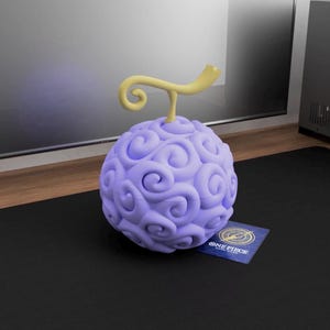 Könnte beinhalten: Eine hellviolette, kugelförmige Frucht mit gelbem Stiel und Wirbeln, die einer Teufelsfrucht ähnelt. Eine kleine Karte mit dem Text "ONE PIECE" befindet sich neben der Frucht. Die Frucht liegt auf einer schwarzen Oberfläche.