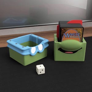 May include: Two green and blue card storage boxes with cartoon-like faces. One box has a blue rim and white eye shapes. The other box has a smiling face. A white die with black dots sits nearby. The boxes hold playing cards.