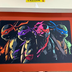 Puede incluir: Alfombrilla de ratón rectangular negra con las Tortugas Ninja. Las cuatro tortugas están representadas en un estilo de cómic, cada una con su máscara de color: rojo, morado, rosa y azul. Salpicaduras de color acentúan el fondo.