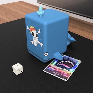 May include: A blue whale-shaped card holder with a skull and crossbones graphic. A trading card is partially inserted into the whale's mouth. A white die is on the black surface.