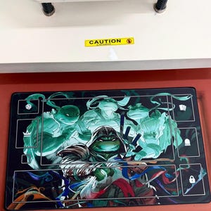 May include: A rectangular black playmat featuring the Teenage Mutant Ninja Turtles in a dynamic, action-packed illustration. The design showcases the turtles in shades of green and blue, with a comic book style. The playmat has white bordered zones and the word "CAUTION" is visible.
