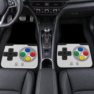 May include: A pair of car floor mats featuring a light gray design modeled after a retro video game controller. Each mat displays a black directional pad, four colored action buttons labeled A, B, X, and Y, and small circles marked Select and Start.