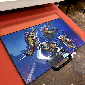May include: A rectangular mouse mat featuring the Teenage Mutant Ninja Turtles in action poses against a cityscape backdrop. The mat has a black border and a black handle.