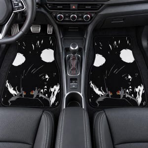 May include: A pair of black car floor mats featuring a graphic illustration of an anime character with spiky hair and glowing red eyes. The design includes white circular shapes and abstract white streaks against a solid black background.
