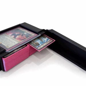 May include: Black card storage box, open to reveal a deck of pink-edged cards and several trading cards. The box is designed to hold and protect collectible cards, with a sliding lid for easy access.
