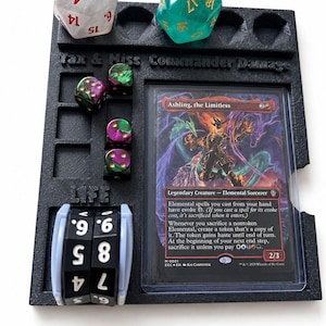 May include: A black plastic Commander Damage and Life counter. The holder has slots for dice and a card. Includes a clear d20, a clear d6, and several purple and green dice. The card reads "Ashling, the Limitless". The text "Tax & Hiss Commander Damage" is visible.