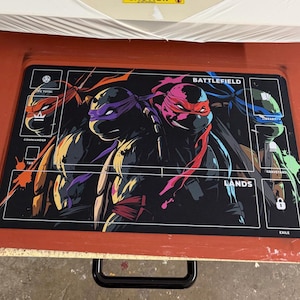 May include: A rectangular black playmat featuring the Teenage Mutant Ninja Turtles. The mat has sections labelled "Commander", "Battlefield", "Lands", and "Exile". The turtles are depicted in their signature colours: red, purple, blue, and orange.