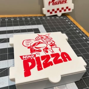 May include: White Ninja Pizza box with red graphics. The box features the words "NINJA PIZZA" in bold red letters, along with a cartoon image of a ninja chef. A second box with the words "Pizza Planet" is visible in the background.