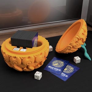 Könnte beinhalten: Orangener, fruchtförmiger Karten- und Würfelhalter mit einem Stapel schwarzer Karten im Inneren. Zwei "One Piece Card Game" Karten und drei weiße Würfel befinden sich neben dem Halter. Der Halter ist geöffnet und gibt den Innenraum frei.