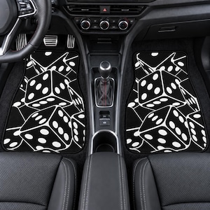 May include: A pair of black car floor mats featuring a repeating white graphic pattern of six-sided dice. The mats are positioned on the floor of a vehicle interior, covering the footwell areas on both the driver and passenger sides of the console.