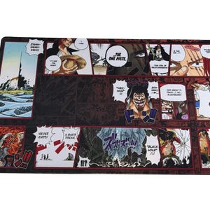 May include: A rectangular black playmat featuring panels of manga artwork with red borders. The panels depict various characters and scenes, including text bubbles and action lines. The text includes "THE ONE PIECE" and "BLACK HOLE!!!".