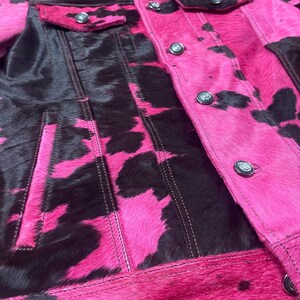 May include: A jacket featuring a vibrant hot pink and black cow print pattern with a textured hair-on-hide finish. The garment includes a classic collar, long sleeves, two chest pockets with button closures, and a front button-down placket.