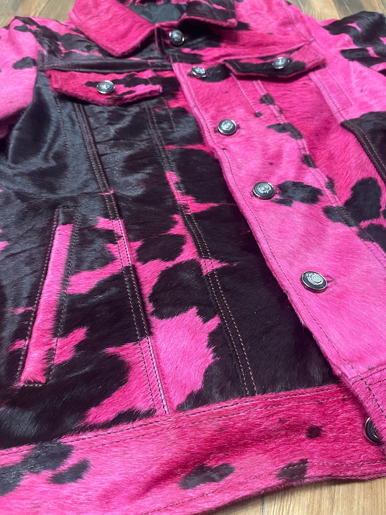 May include: A jacket featuring a vibrant hot pink and black cow print pattern with a textured hair-on-hide finish. The garment includes a classic collar, long sleeves, two chest pockets with button closures, and a front button-down placket.