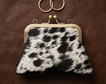 May include: A small, black and white cowhide coin purse with a brass clasp and two gold hoop earrings. The purse has a unique spotted pattern and is set against a brown leather background.