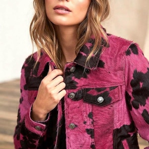 May include: A long-sleeve jacket featuring a vibrant pink and black cow print pattern with a textured hair-on-hide finish. The garment includes a classic collar, metal button closures down the front, buttoned cuffs, and two chest pockets with flaps.