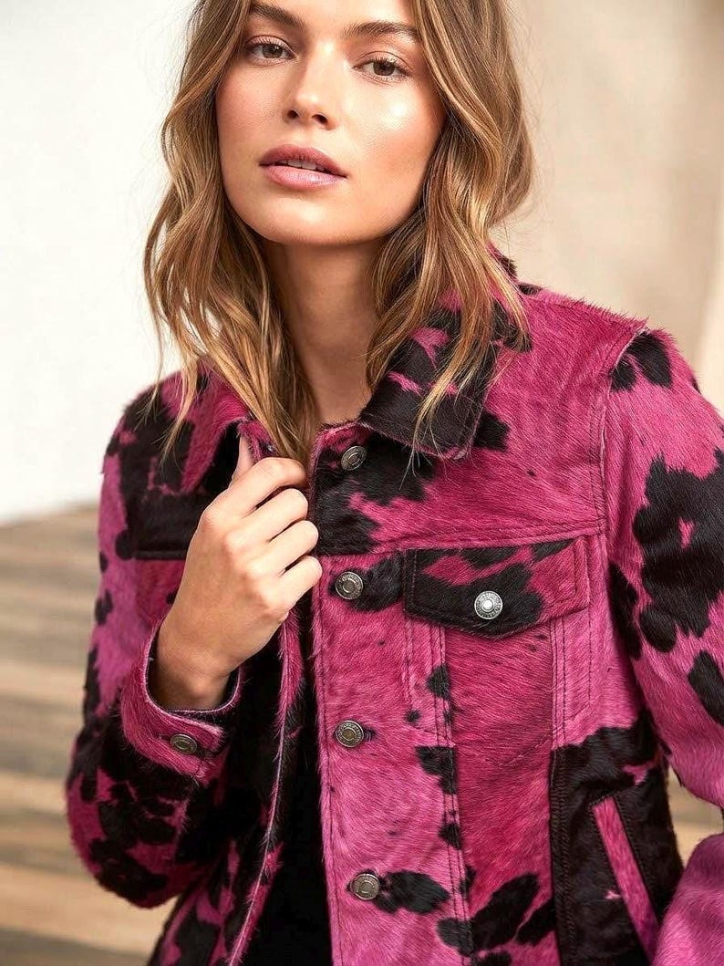 May include: A long-sleeve jacket featuring a vibrant pink and black cow print pattern with a textured hair-on-hide finish. The garment includes a classic collar, metal button closures down the front, buttoned cuffs, and two chest pockets with flaps.
