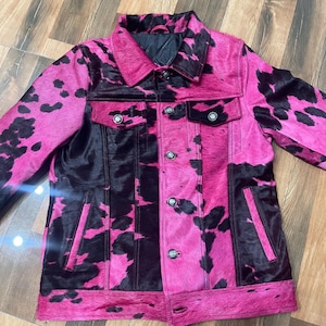May include: A long-sleeve jacket featuring a vibrant hot pink and black cow print pattern. The garment includes a classic collar, a front button closure with metallic buttons, two chest pockets with buttoned flaps, and two vertical side pockets.