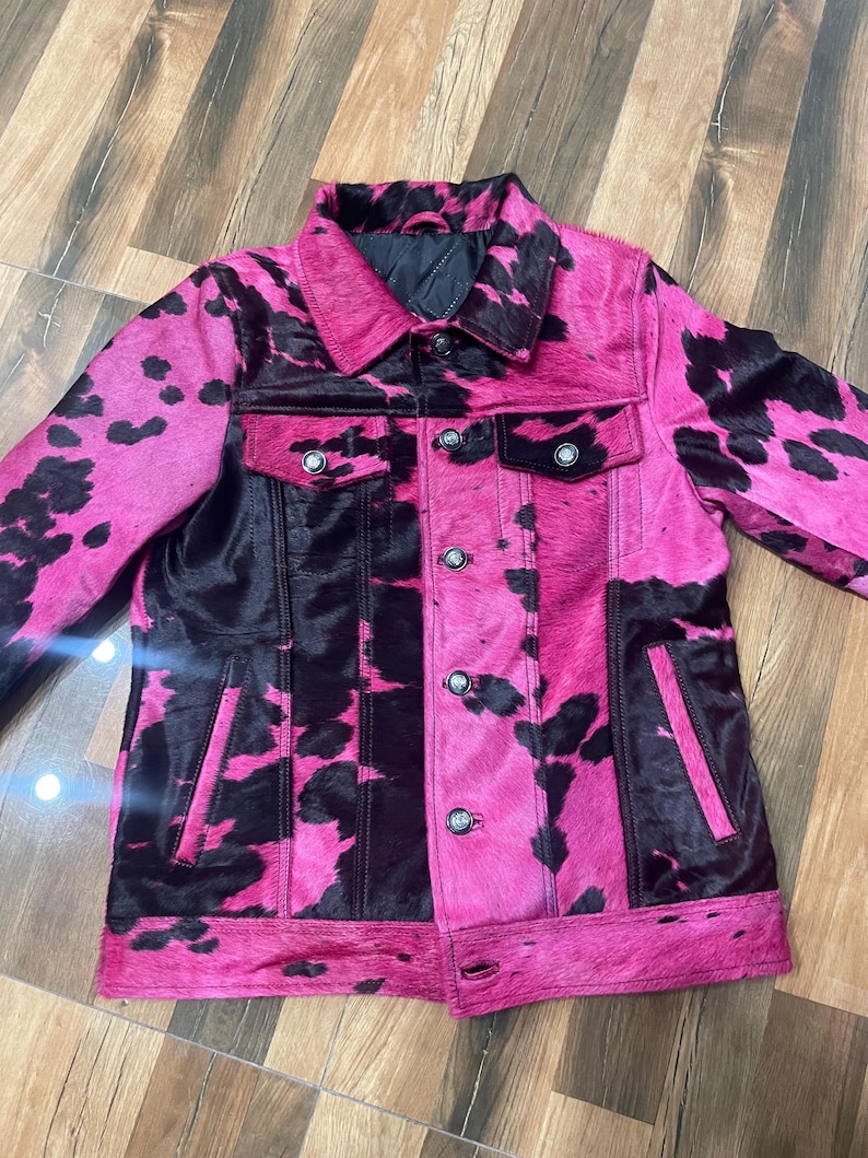 May include: A long-sleeve jacket featuring a vibrant hot pink and black cow print pattern. The garment includes a classic collar, a front button closure with metallic buttons, two chest pockets with buttoned flaps, and two vertical side pockets.