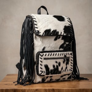 May include: A backpack featuring a black and white cow print pattern with long black fringe detailing on the sides. The bag includes a top flap with a zip closure, a front zipped pocket, and black leather-like trim with decorative stitching.
