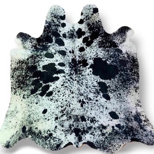 May include: A cowhide rug featuring an irregular, organic shape with a natural pattern of black and white spots. The surface displays a dense concentration of small black speckles transitioning into larger, solid black patches against a white base.
