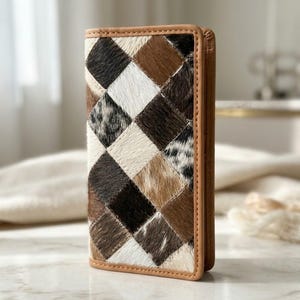 Puede incluir: Una cartera rectangular con un diseño de patchwork de cuadrados de piel de vaca en tonos marrón, negro, blanco y gris. La cartera está enmarcada con un borde de cuero color canela liso a lo largo de los lados, creando un aspecto estructurado.