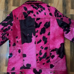 May include: A jacket featuring a vibrant hot pink and black cow print pattern. The garment has a collared neckline, long sleeves, and a button-down front closure. The texture appears to be a short-haired hide, with visible vertical seams and metallic buttons at the hem.