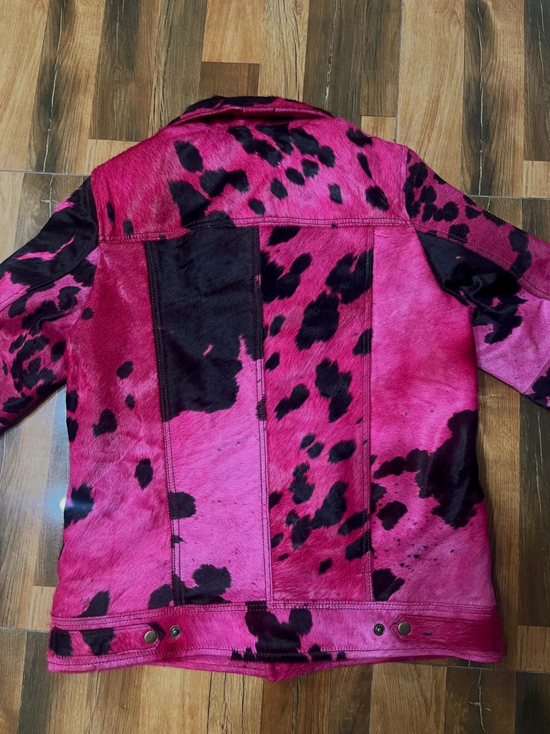 May include: A jacket featuring a vibrant hot pink and black cow print pattern. The garment has a collared neckline, long sleeves, and a button-down front closure. The texture appears to be a short-haired hide, with visible vertical seams and metallic buttons at the hem.