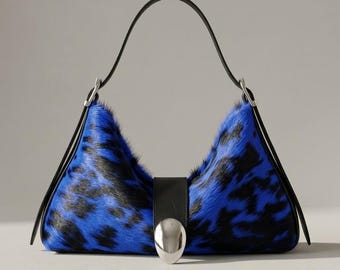 May include: A crescent-shaped shoulder bag featuring a vibrant blue and black animal print fur texture. The bag includes a smooth black leather strap and a central black leather tab secured by a large, polished silver-tone metal oval clasp.