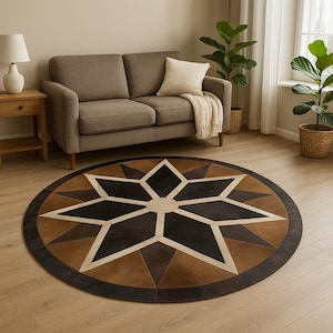 May include: A round area rug with a starburst design. The rug features a central star pattern in black, cream, and brown tones, surrounded by a border of darker brown. The rug is placed on a light wood floor.
