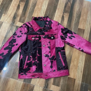 May include: A long-sleeve jacket featuring a vibrant hot pink and black cow print pattern. The garment includes a classic collar, a front button-down closure with metallic buttons, two chest pockets with button flaps, and two vertical side pockets.