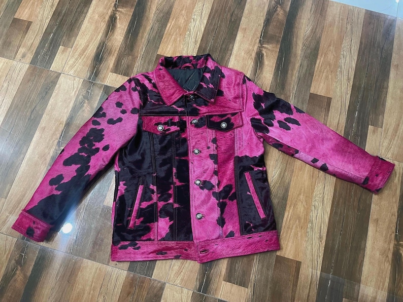 May include: A long-sleeve jacket featuring a vibrant hot pink and black cow print pattern. The garment includes a classic collar, a front button-down closure with metallic buttons, two chest pockets with button flaps, and two vertical side pockets.