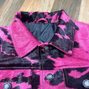 May include: A jacket featuring a vibrant hot pink and black cow print pattern with a textured hair-on-hide finish. The garment includes a classic collar, a black quilted interior lining, and dark metallic buttons on the front placket and chest pockets.
