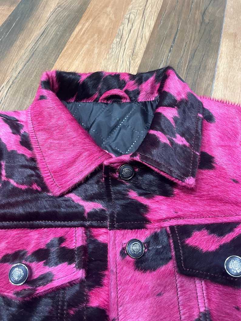 May include: A jacket featuring a vibrant hot pink and black cow print pattern with a textured hair-on-hide finish. The garment includes a classic collar, a black quilted interior lining, and dark metallic buttons on the front placket and chest pockets.