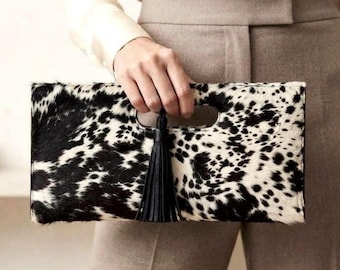 May include: A rectangular clutch bag featuring a black and white cowhide pattern with a textured fur finish. The bag includes a central cutout handle and a long, black leather tassel attached to the top edge of the handle opening.