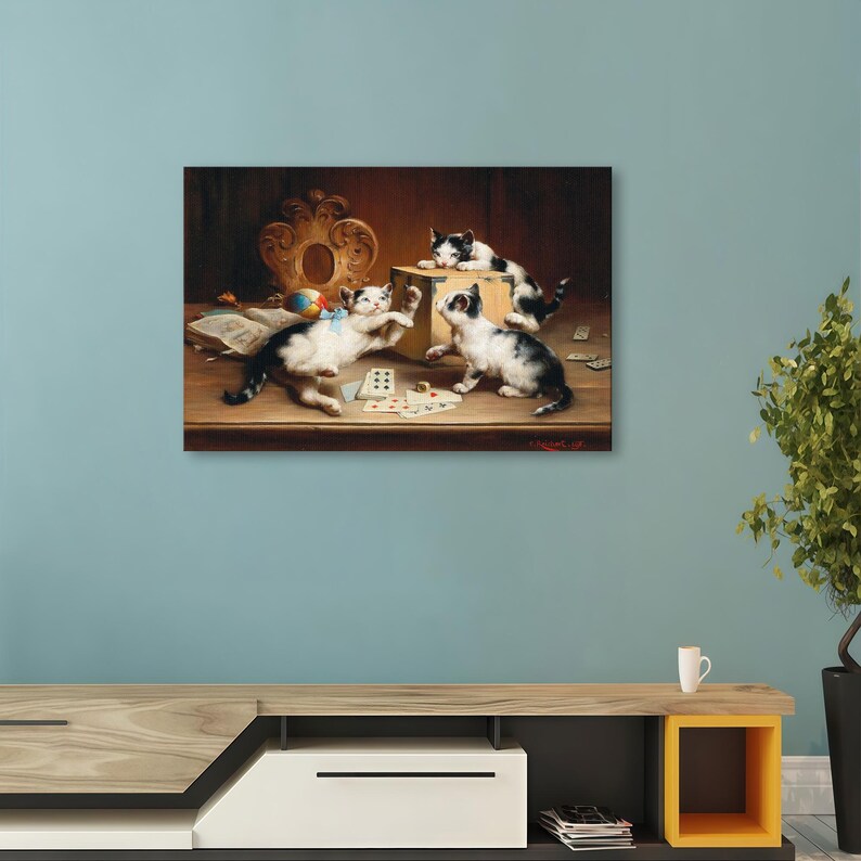 May include: A framed painting showing three kittens, black and white, at play with a wooden box, cards, and a ball. The kittens are the focus, set against a warm, detailed background with a decorative frame.