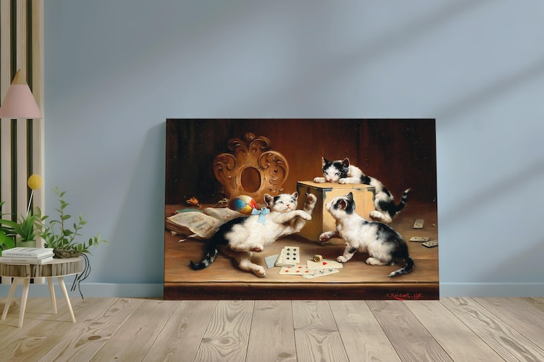 May include: A framed painting showing three kittens at play. Two black and white kittens are interacting with a wooden box, while another kitten reaches for a ball. Playing cards and a book are also on the table.
