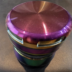 Peut inclure: Un broyeur cylindrique avec une finition métallique. Le dessus est d'un violet profond avec un motif radial, passant à une bande violette et dorée vibrante. La base présente un effet irisé arc-en-ciel.