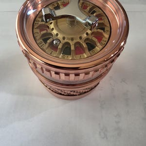 Puede incluir: Un molinillo cilíndrico de color cobre con un diseño de ruleta en la tapa superior. La tapa tiene una ventana circular transparente, un girador plateado de tres puntas y una pequeña bola de metal sobre la superficie dorada numerada.