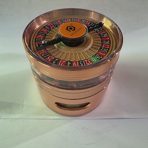 Peut inclure: Un grinder en forme de roue de roulette, de couleur or rose, avec un dessus transparent. La roue comporte des chiffres et un disque central orange avec le mot "PALPITATE". Le grinder est de forme cylindrique.