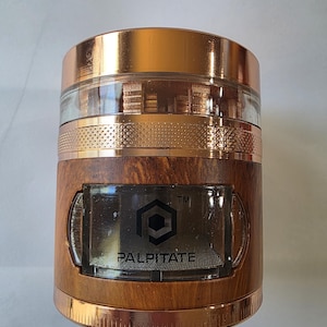 Peut inclure: Un broyeur cylindrique avec un dessus et un dessous de couleur cuivre, et une section centrale imitation bois. Le broyeur a une section en verre transparent et une fenêtre rectangulaire avec le mot "PALPITATE" et un motif géométrique.