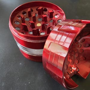 Peut inclure: Un broyeur en métal rouge avec une bande argentée. Le broyeur est ouvert, révélant l'intérieur avec de multiples trous. La partie supérieure détachée est également rouge, avec un motif texturé. Le broyeur repose sur une surface sombre.