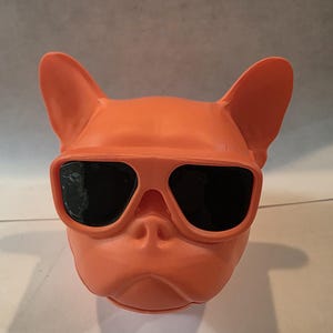 Peut inclure: Figurine de tête de bouledogue orange portant des lunettes de soleil à verres noirs. La figurine a de grandes oreilles et un museau détaillé. Les lunettes de soleil sont orange et assorties à la couleur de la tête du bouledogue. Objet décoratif amusant et original.