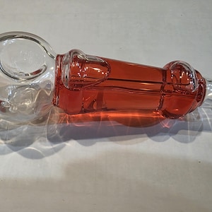 May include: A clear glass pipe with a bulbous end and a red liquid filling the central chamber. The pipe has a rounded mouthpiece and a flared end. The glass is smooth and reflects light.