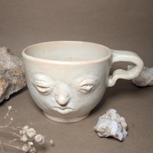 Könnte beinhalten: Cremefarbene Keramiktasse mit einem geformten Gesichtsdesign. Die Tasse hat geschlossene Augen, eine Nase und einen Mund. Sie hat einen Henkel und steht auf einem neutralen Hintergrund mit Steinen und Trockenblumen.