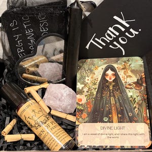 Puede incluir: Una caja negra llena de artículos espirituales. Incluye un frasco de perfume, una vela negra, una tarjeta con el texto "Thank you", una tarjeta con el texto "DIVINE LIGHT", una bolsa de malla negra con el texto "ENERGY IS MAGNETIC DESIRE", cristales y otros artículos.