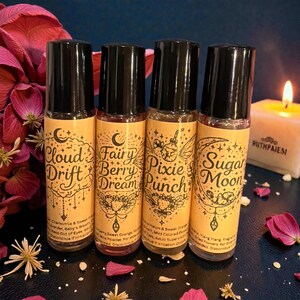 May include: Four roll-on perfume bottles with black caps and labels featuring whimsical designs and text. The labels read "Cloud Drift," "Fairy Berry Dream," "Pixie Punch," and "Sugar Moon." A lit candle and flower petals are in the background.